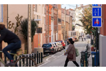 Rue avec présence d'une piétonne et d'un cycliste accompagnés de panneaux indiquant la présence d'une vélorue